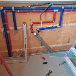 Ściana z drewnianych bali z zamontowanymi rurami wodnymi w izolacji czerwonej i niebieskiej, poziomica przy ścianie, widoczne narzędzia i fragment drabiny.