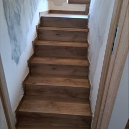 DARIUSZ KUTARNIA DARKFLIZ - Wykończone schody drewniane wewnątrz budynku, prowadzące na górę. Ściany częściowo przygotowane do malowania, widoczne niedoskonałości. Naturalne światło wpada przez okno.