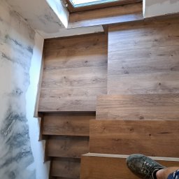 DARIUSZ KUTARNIA DARKFLIZ - Wykończone drewnopodobne schody z oknem na podeście, kontrastujące z dekoracyjną ścianą o efekcie marmuru. Widoczny fragment buta osoby wykonującej pracę.