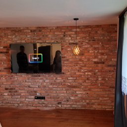 DARIUSZ KUTARNIA DARKFLIZ - Salon z ceglaną ścianą, na której wisi telewizor. Po bokach lampy, a z prawej strony okno z firanką. Na pierwszym planie kobieta w sukience w kropki.