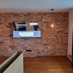 DARIUSZ KUTARNIA DARKFLIZ - Ceglana ściana z wiszącym telewizorem i lampami. Widoczny fragment podłogi i okno z zasłonami. Dekoracyjne wykończenie wnętrza z elementami oświetlenia.