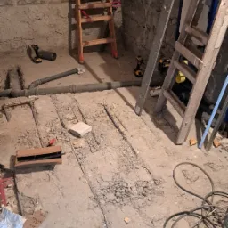 Instalacja ogrzewania podłogowego w surowym pomieszczeniu z kamienną ścianą. Widoczne rury, drabiny i narzędzia. Prace hydrauliczne w trakcie realizacji.