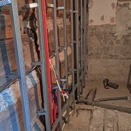 DARIUSZ KUTARNIA DARKFLIZ - Stelaż z profili metalowych przy drewnianej ścianie, czerwone rury instalacji, surowy stan, przygotowanie do wykończenia wnętrza. Widoczne narzędzia i elementy instalacji.