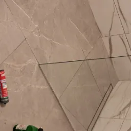 Wykończona łazienka z beżowymi płytkami imitującymi marmur, kabina prysznicowa ze szklaną ścianką, widoczny odpływ liniowy i akcesoria glazurnicze.