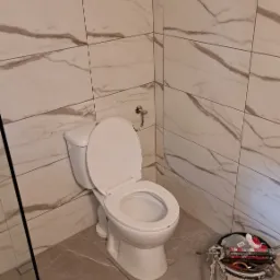 Biała toaleta z otwartą klapą w wykończonej łazience. Marmurowe płytki na ścianach i podłodze. Widoczne narzędzia pracy i butelka. Ukończona realizacja.