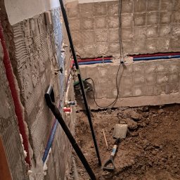 DARIUSZ KUTARNIA DARKFLIZ - Przygotowanie do remontu: ściany z resztkami zaprawy, rury instalacyjne, łopata i ziemia na podłodze. Widoczne prace instalacyjne w pomieszczeniu.