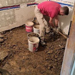 DARIUSZ KUTARNIA DARKFLIZ - Demontaż posadzki w łazience: mężczyzna w czerwonej koszulce usuwa ziemię łopatą, widoczne ściany z pozostałościami płytek i rury instalacji.