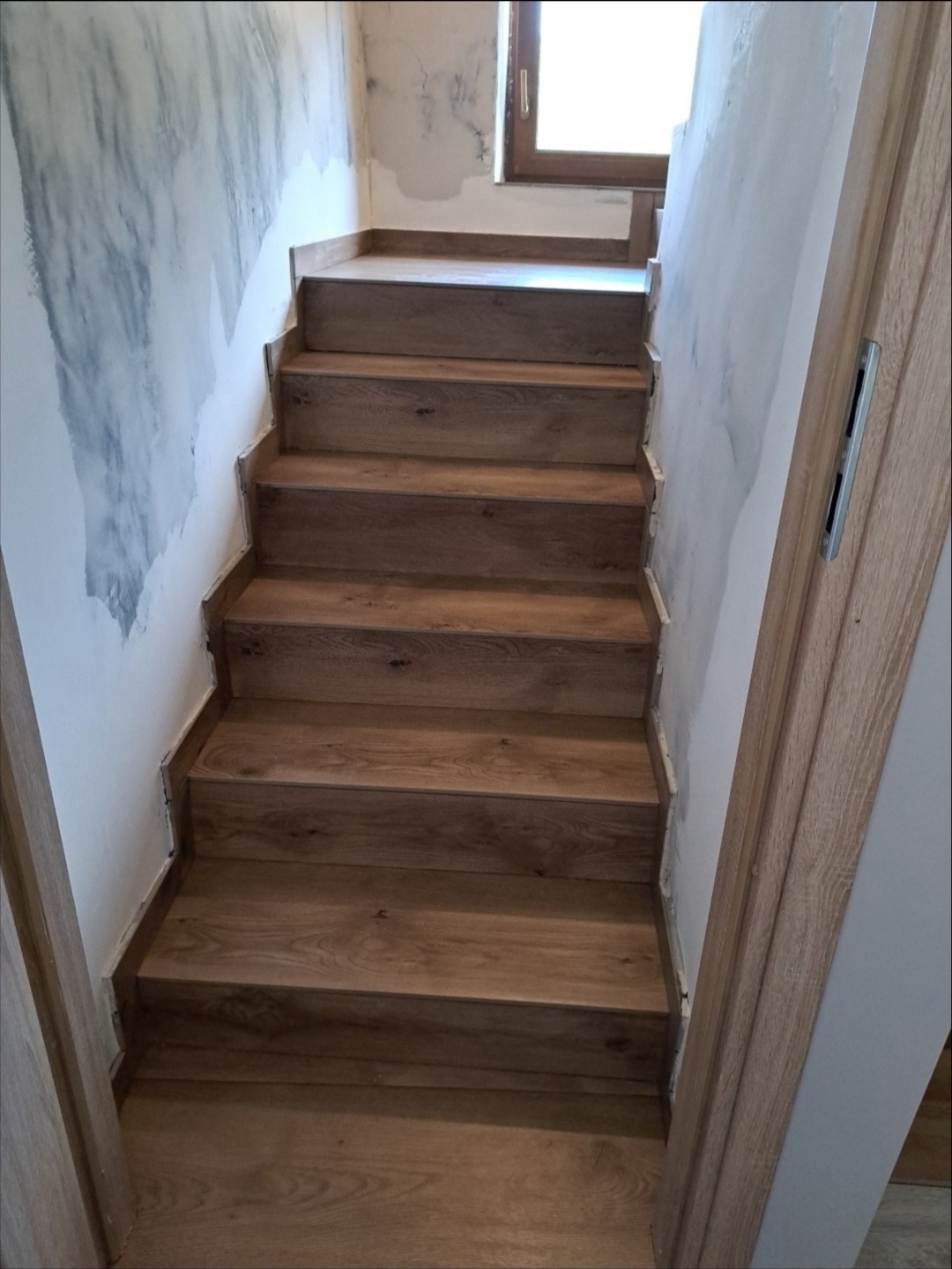 Wykończone schody drewniane wewnątrz budynku, prowadzące na górę. Ściany częściowo przygotowane do malowania, widoczne niedoskonałości. Naturalne światło wpada przez okno.