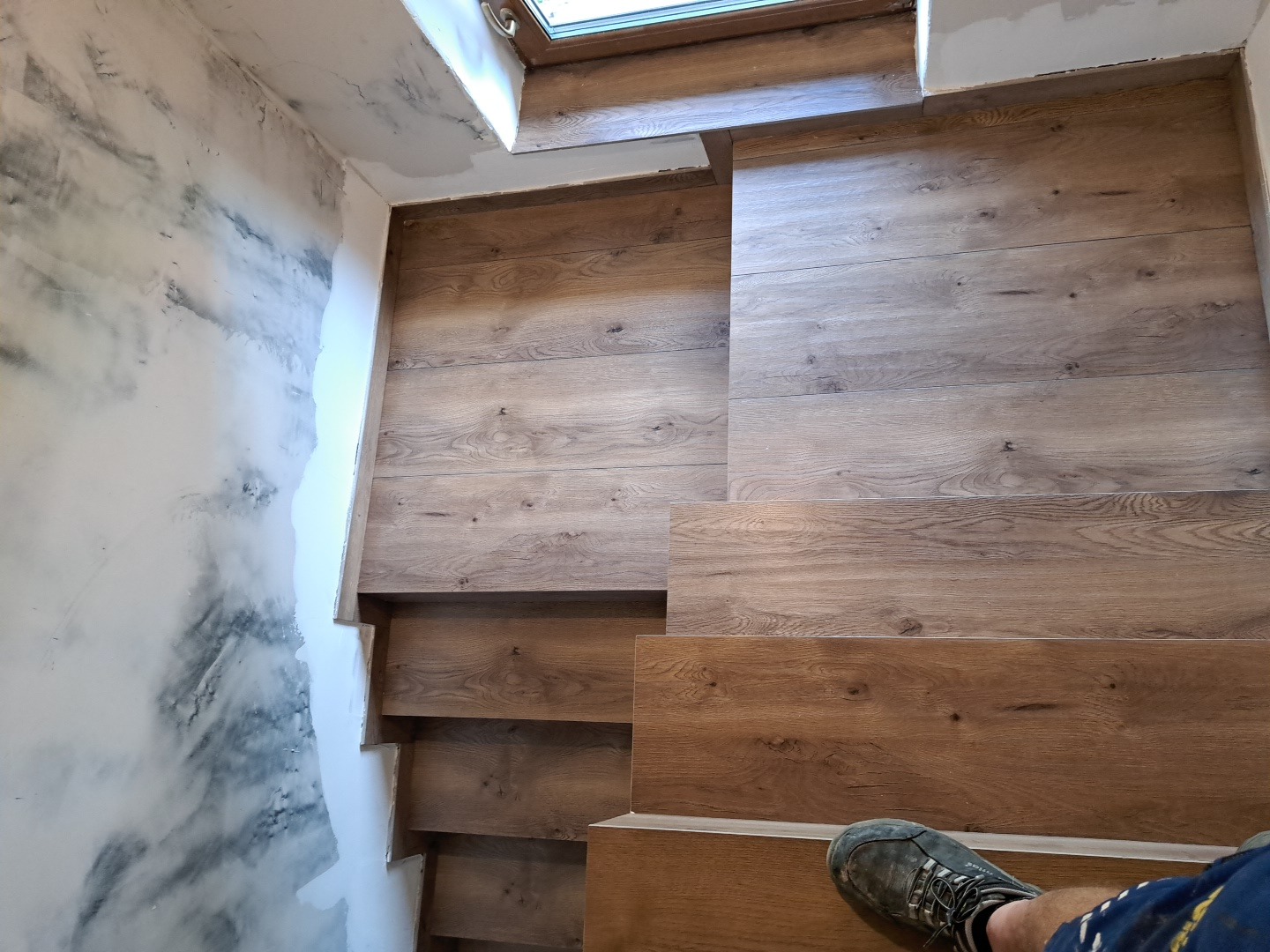 Wykończone drewnopodobne schody z oknem na podeście, kontrastujące z dekoracyjną ścianą o efekcie marmuru. Widoczny fragment buta osoby wykonującej pracę.