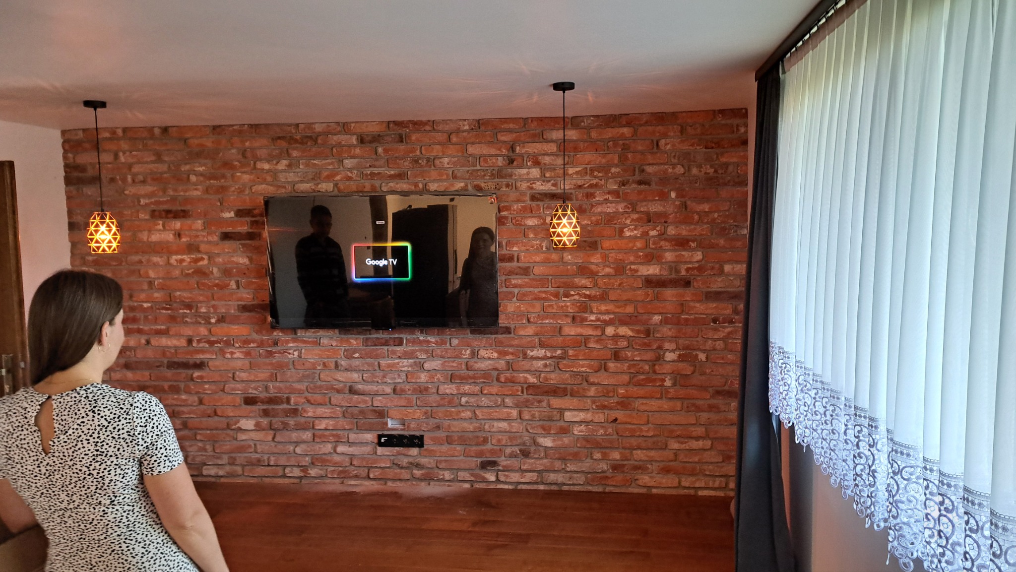 Salon z ceglaną ścianą, na której wisi telewizor. Po bokach lampy, a z prawej strony okno z firanką. Na pierwszym planie kobieta w sukience w kropki.