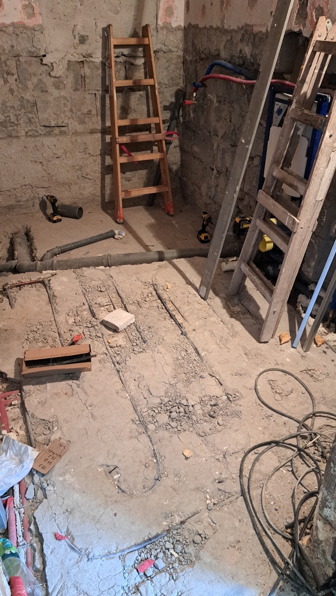 Instalacja ogrzewania podłogowego w surowym pomieszczeniu z kamienną ścianą. Widoczne rury, drabiny i narzędzia. Prace hydrauliczne w trakcie realizacji.