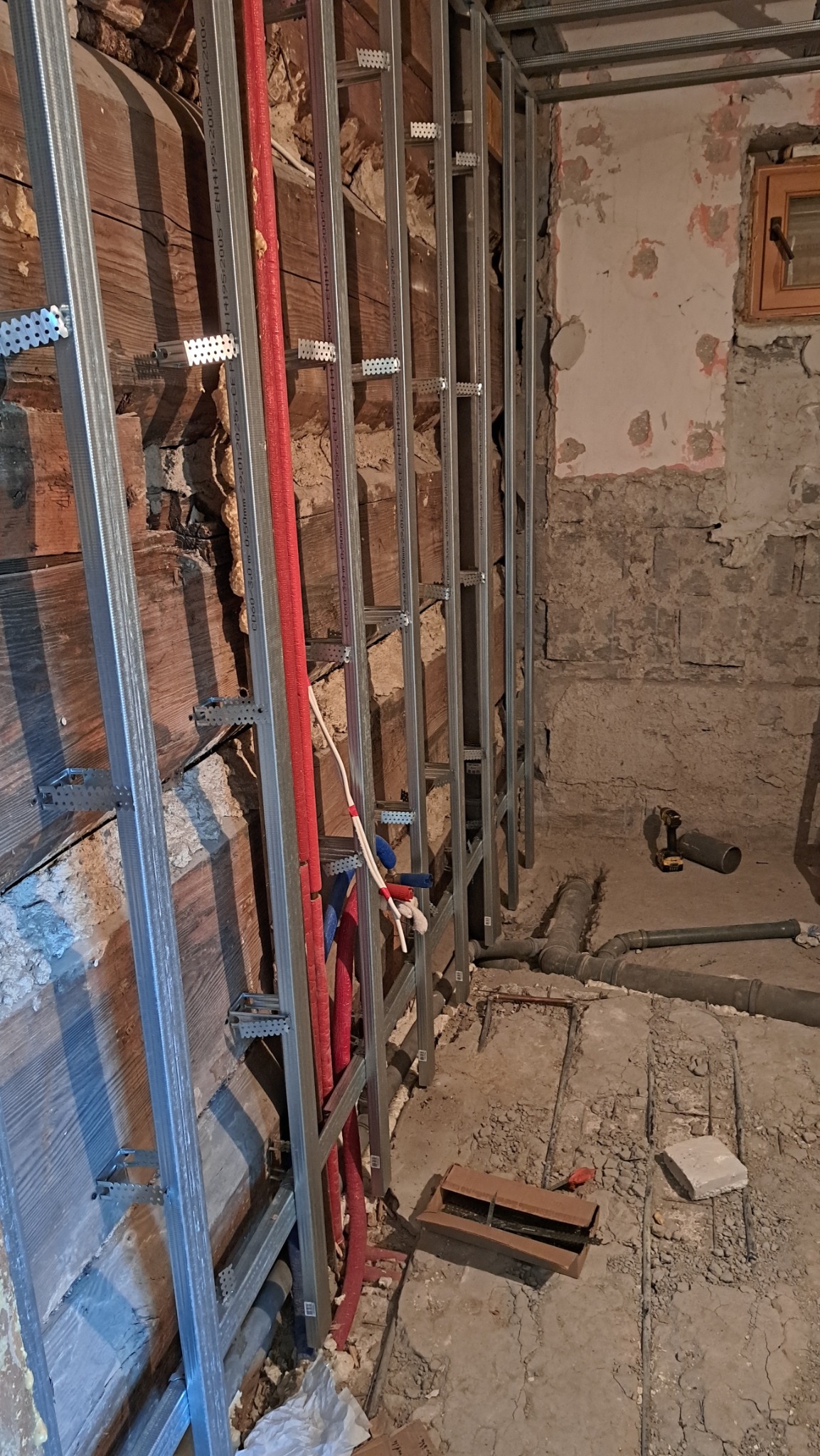 Stelaż z profili metalowych przy drewnianej ścianie, czerwone rury instalacji, surowy stan, przygotowanie do wykończenia wnętrza. Widoczne narzędzia i elementy instalacji.