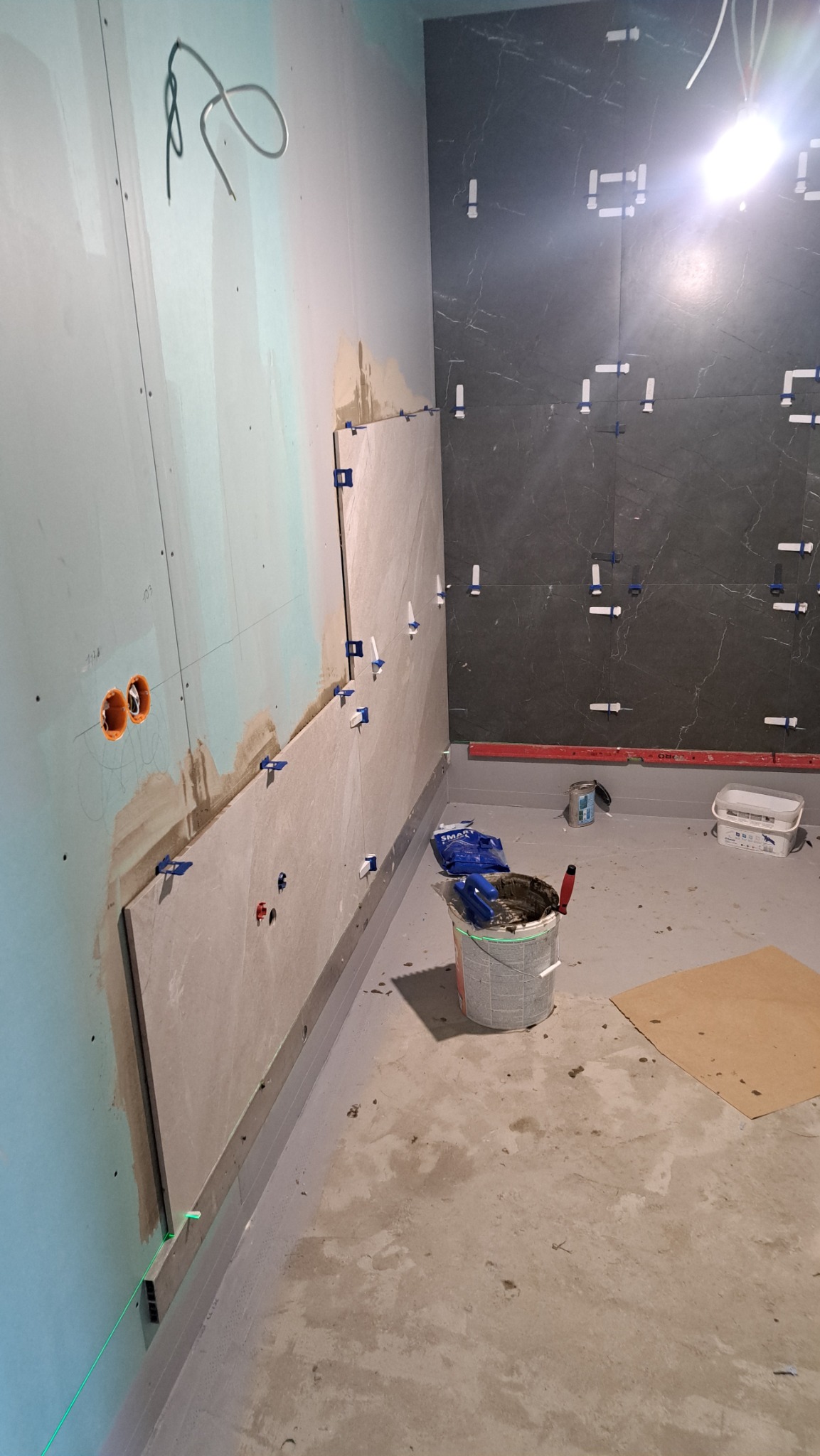 Układanie płytek na ścianie z użyciem poziomicy laserowej i klipsów poziomujących. Widoczny wiadro z zaprawą i narzędzia glazurnicze. Częściowo wykończona ściana w ciemnych płytkach.