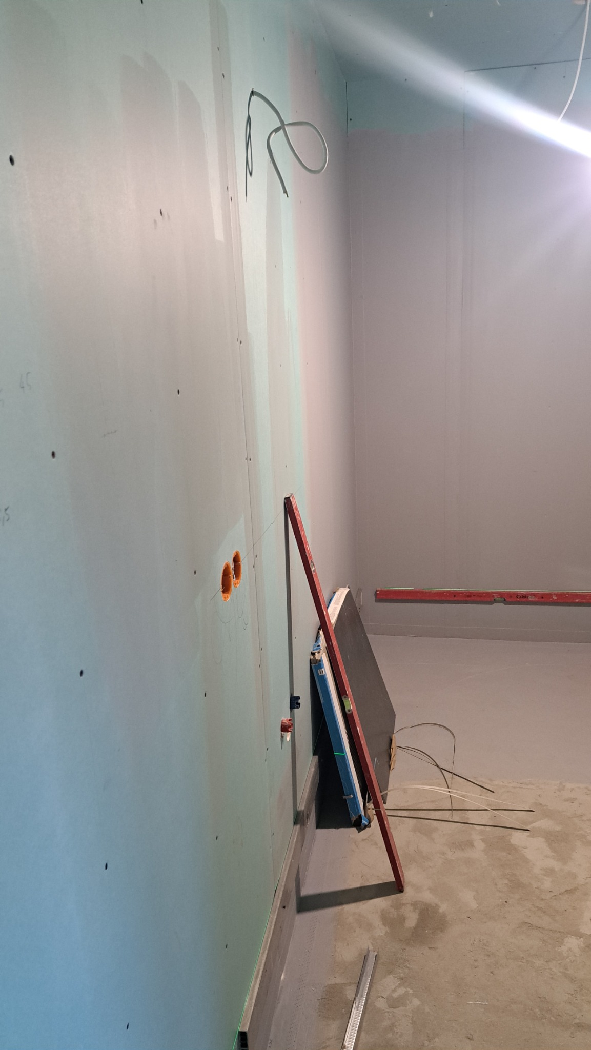 Ściana z płyt gipsowo-kartonowych w trakcie montażu, widoczne profile, otwory na gniazdka, poziomica i przewody. W tle surowy beton i ślady po instalacjach.