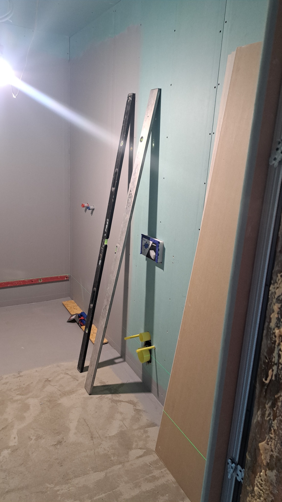 Remont łazienki w trakcie: poziomice opierają się o ścianę z płyt gips-karton, widoczne elementy instalacji hydraulicznej i zielona linia lasera. Surowy beton na podłodze.