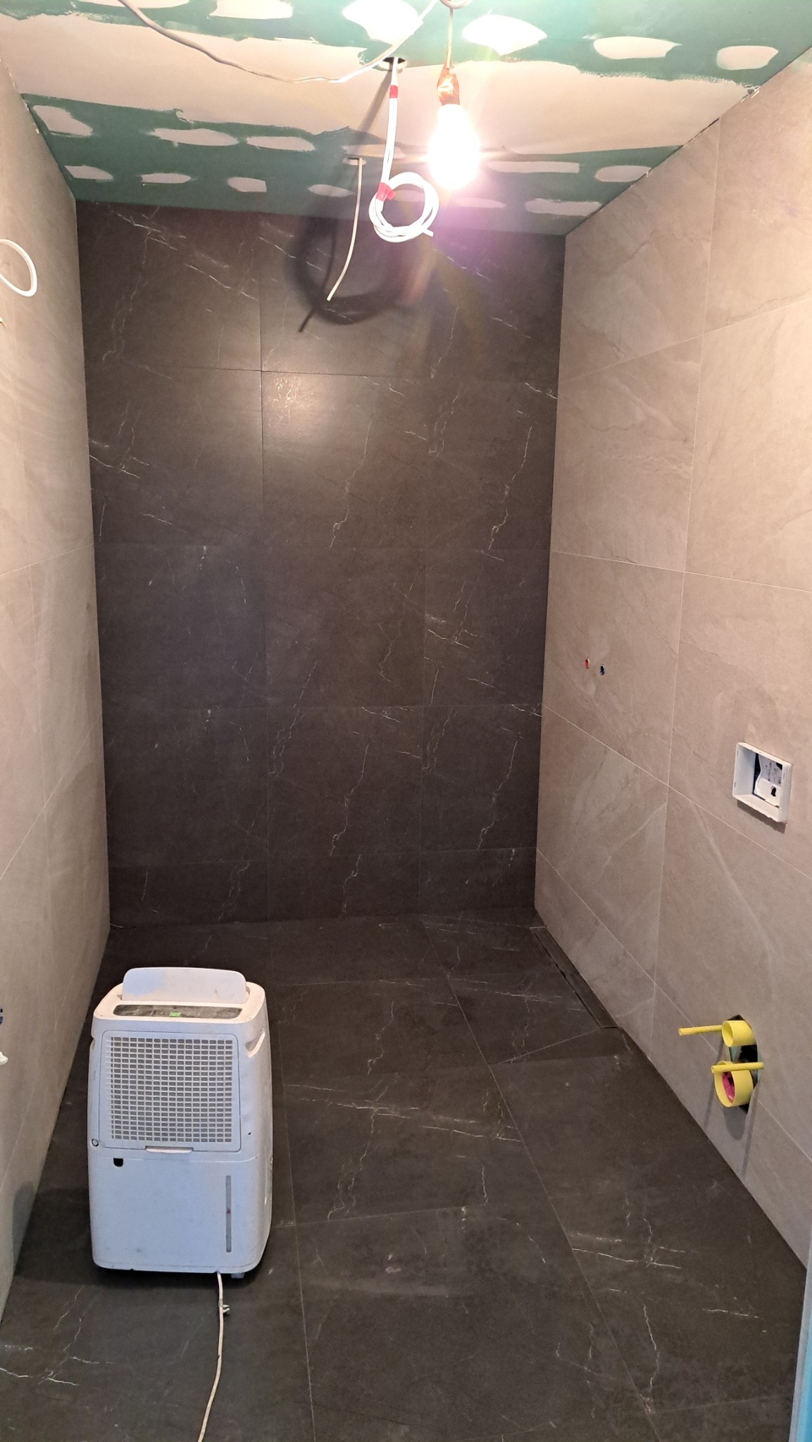 Wykończona łazienka z ciemnymi i jasnymi płytkami imitującymi marmur, widoczne instalacje i osuszacz powietrza w rogu, sufit przygotowany pod montaż płyty.