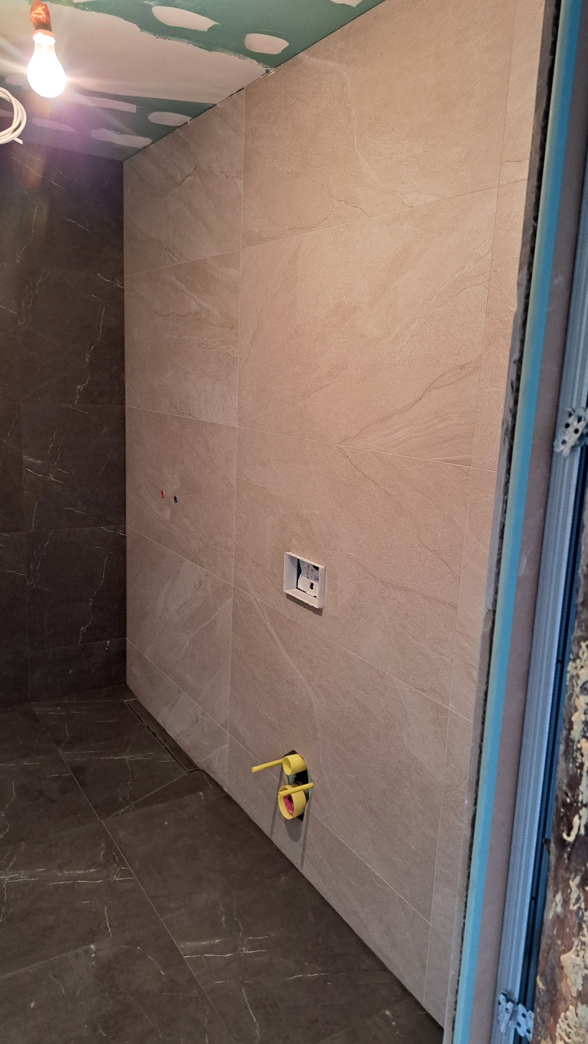 Wykończona łazienka z beżowymi płytkami na ścianach i ciemnymi na podłodze. Widoczne wystające rury i sufit z płyt kartonowo-gipsowych.