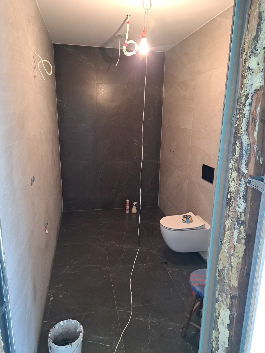 Wykończona łazienka z czarnymi płytkami imitującymi marmur na podłodze i ścianie pod prysznicem. Widoczna toaleta, biała ściana i fragment ościeżnicy z pianką montażową.