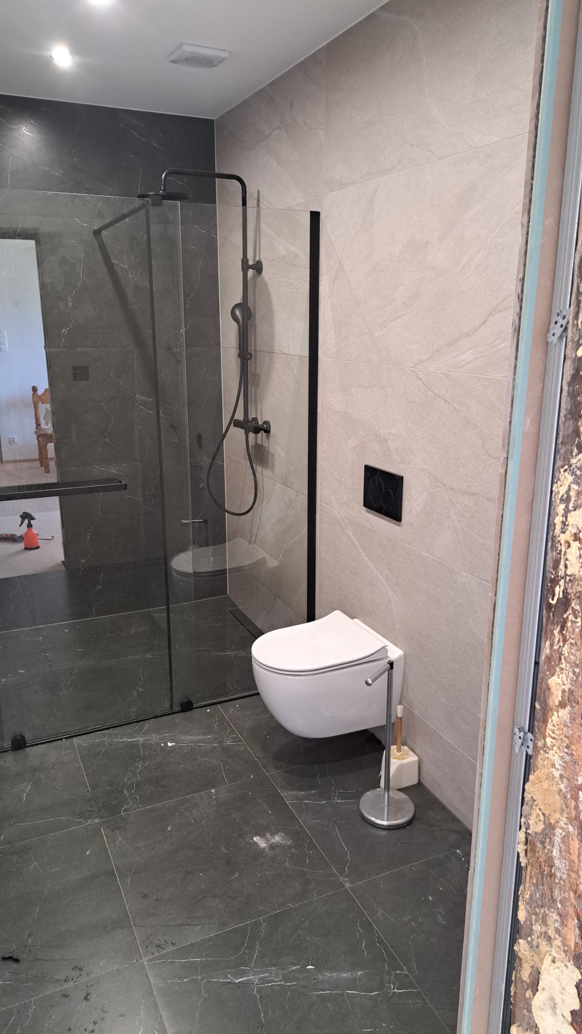 Wykończona łazienka z kabiną prysznicową typu walk-in, wiszącą toaletą i płytkami imitującymi marmur. Widoczne ślady po fugowaniu na podłodze.