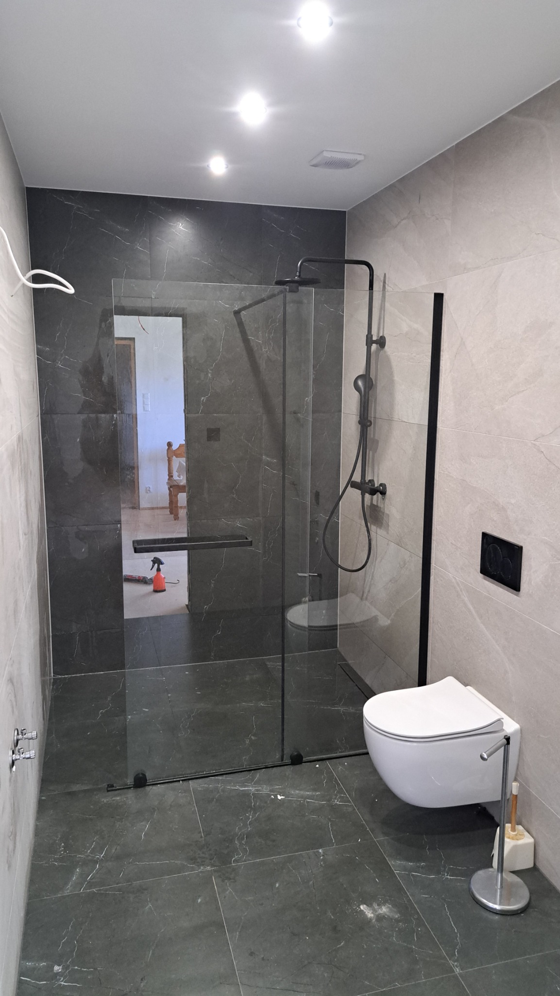 Nowoczesna łazienka z kabiną typu walk-in, czarną armaturą i marmurowymi płytkami. W tle widoczne pomieszczenie z krzesłem, co sugeruje otwarty plan.