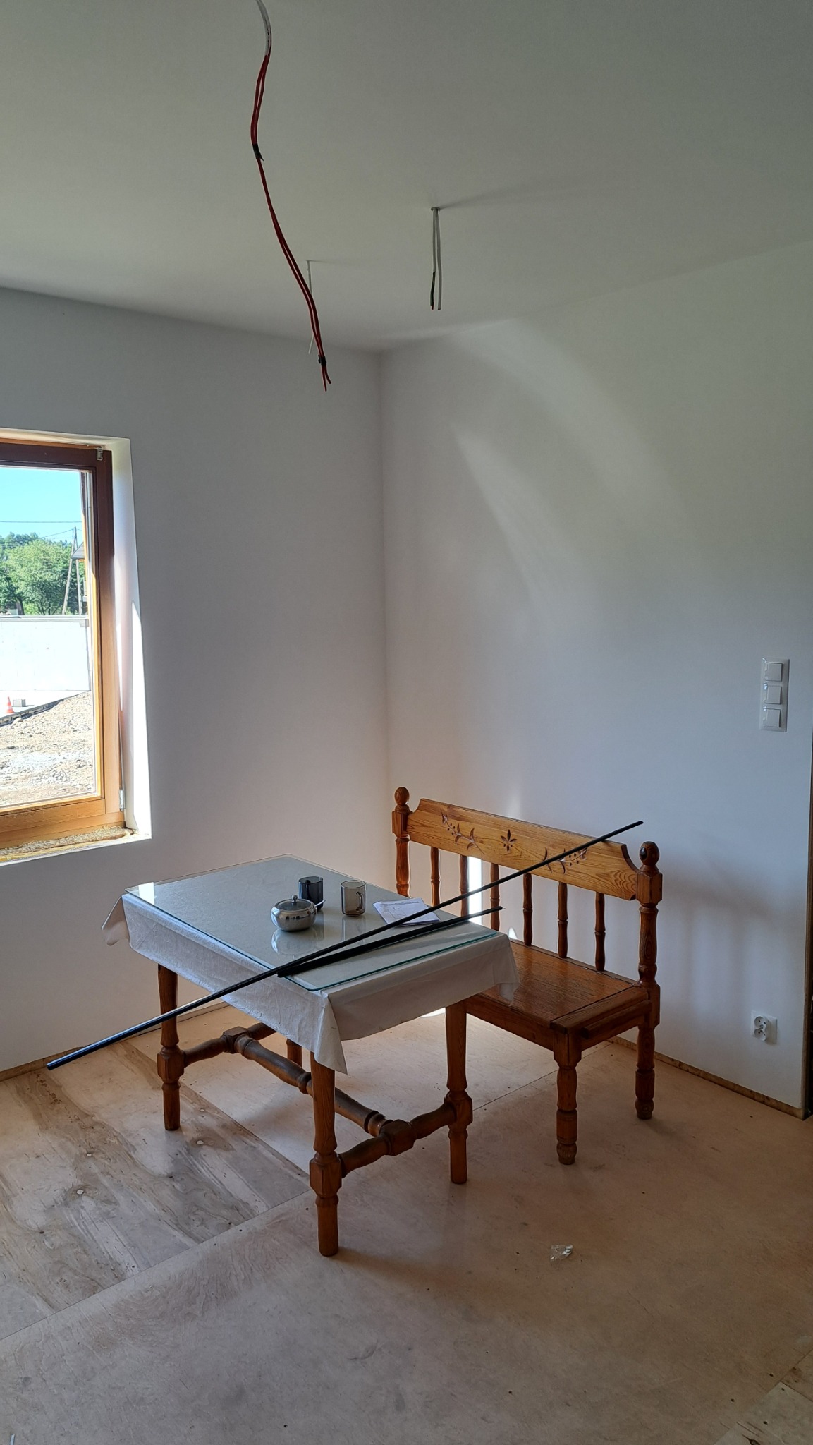 Wnętrze pokoju w trakcie wykańczania: widoczne kable wystające z sufitu, okno z widokiem na zewnątrz, drewniany stół i krzesło. Podłoga w jasnych panelach.