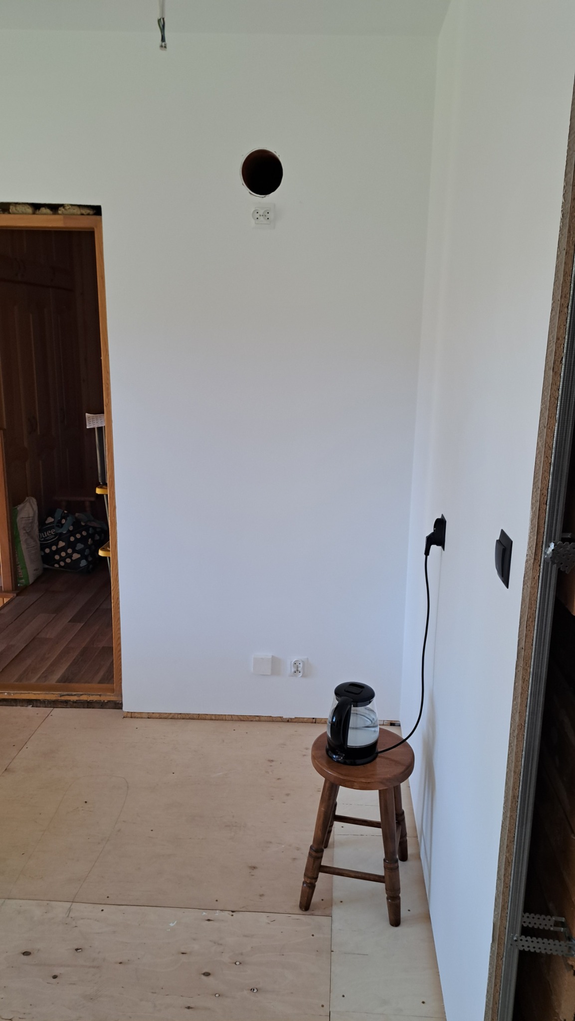 Białe ściany w trakcie prac wykończeniowych, widoczne gniazdka, otwór w ścianie, czajnik na stołku. Podłoga z płyt OSB. Fragment konstrukcji z płyt karton-gips.