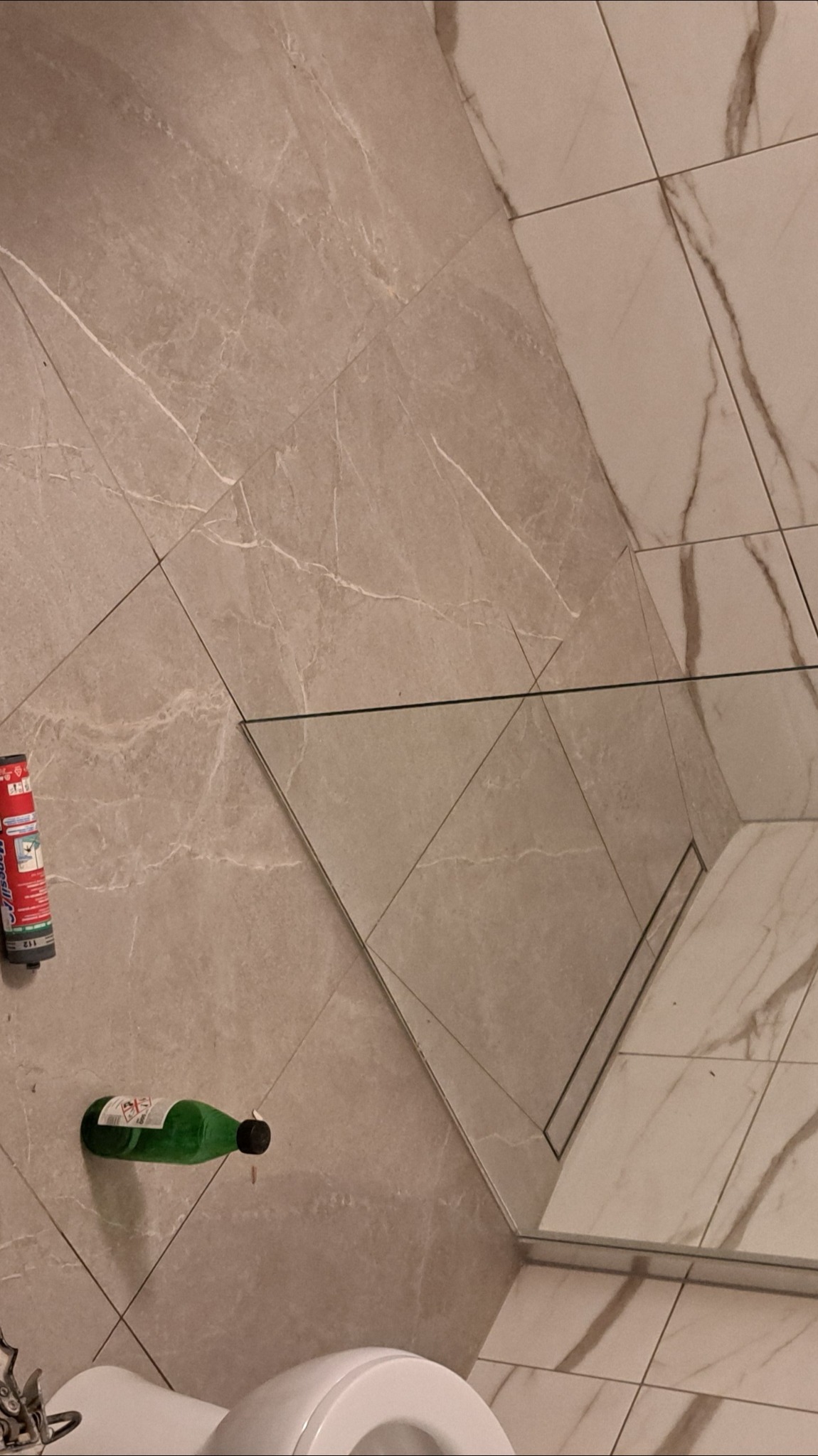 Wykończona łazienka z beżowymi płytkami imitującymi marmur, kabina prysznicowa ze szklaną ścianką, widoczny odpływ liniowy i akcesoria glazurnicze.