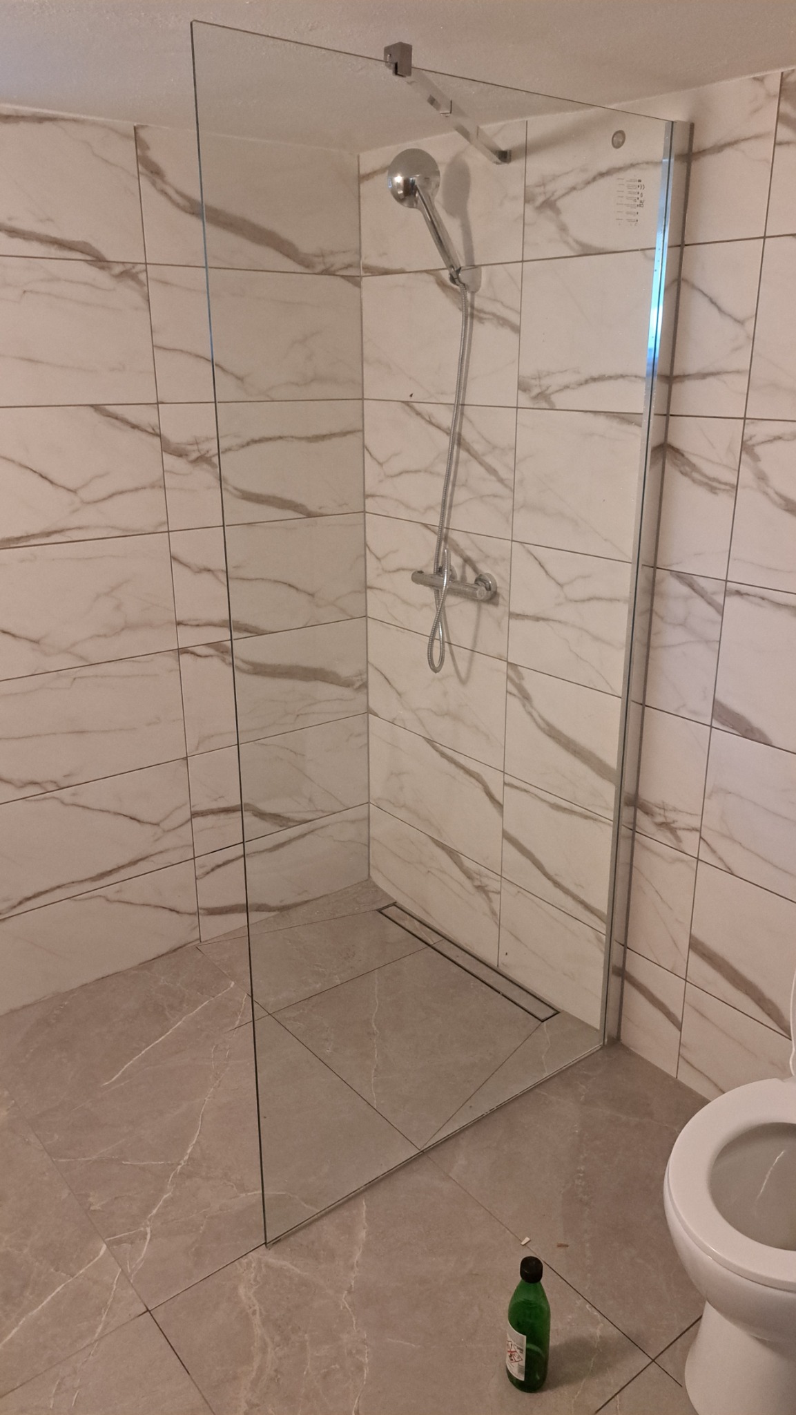 Nowoczesna kabina prysznicowa walk-in z przezroczystego szkła, białe płytki imitujące marmur na ścianach, szary gres na podłodze. Widoczny odpływ liniowy i fragment toalety.