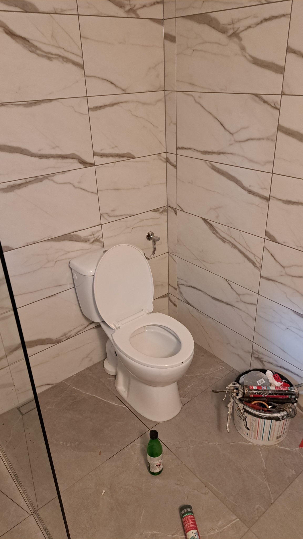 Biała toaleta z otwartą klapą w wykończonej łazience. Marmurowe płytki na ścianach i podłodze. Widoczne narzędzia pracy i butelka. Ukończona realizacja.