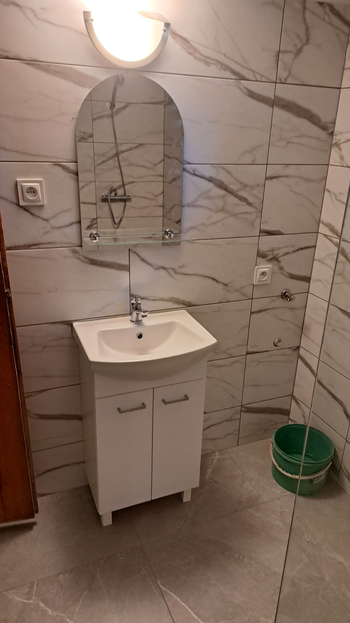 Umywalka z szafką w łazience wykończonej płytkami imitującymi marmur. Nad nią lustro z półką i lampa. Widoczny fragment drzwi i wiadro.