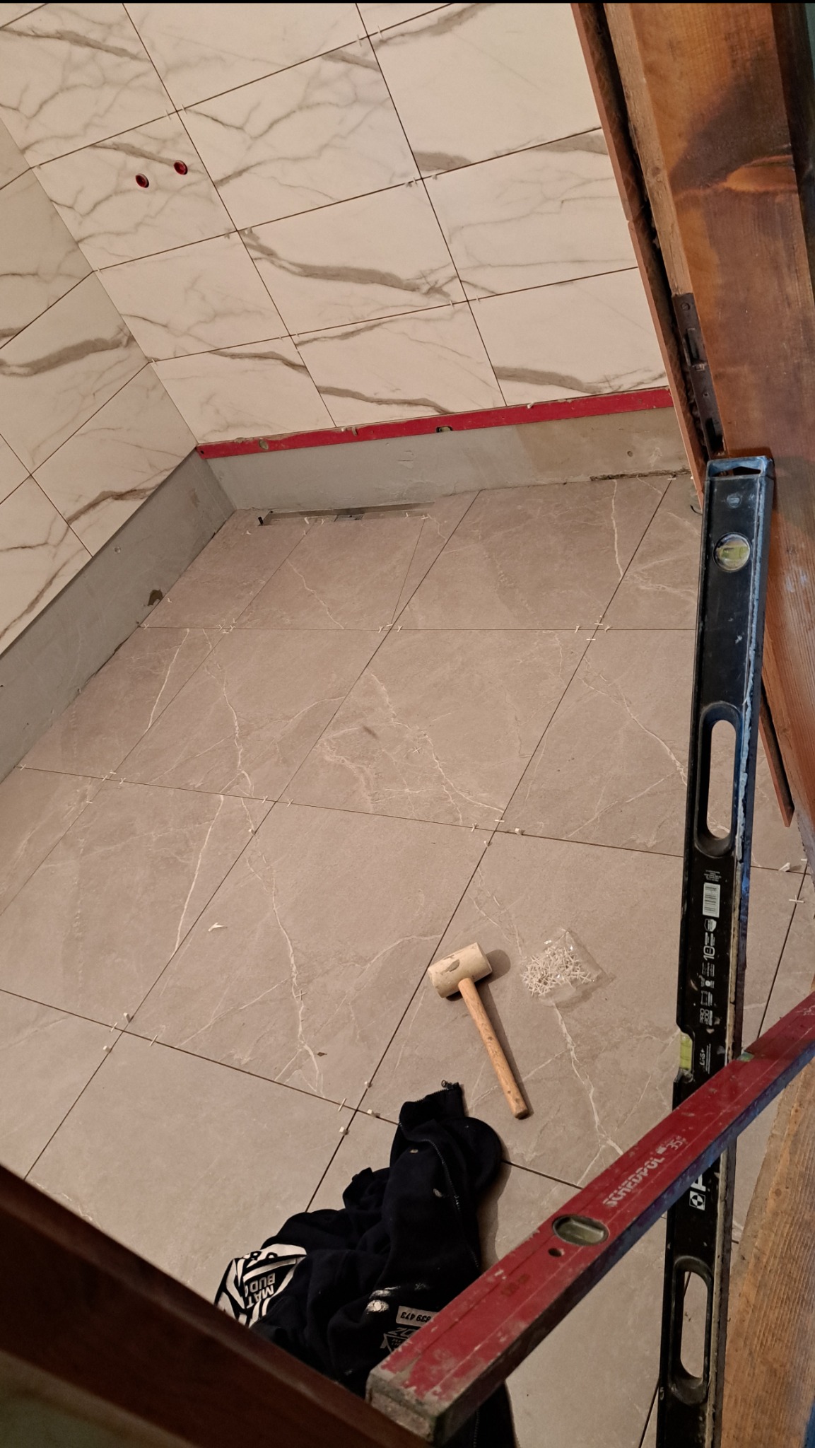 Układanie płytek podłogowych z użyciem poziomicy, młotka i krzyżyków dystansowych. Widoczne płytki imitujące marmur, częściowo ułożone, etap prac wykończeniowych.