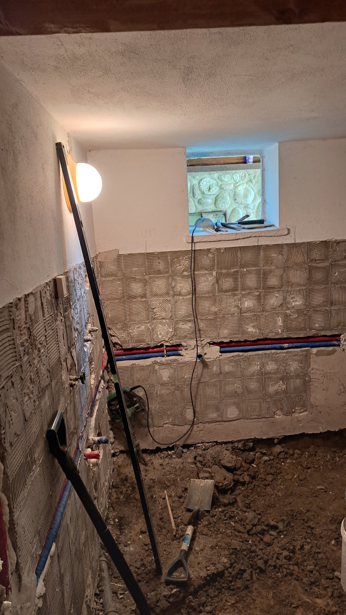 Demontaż płytek w łazience z widocznymi rurami instalacji wodnej. Ściany przygotowane do dalszych prac. Na podłodze ziemia i narzędzia.