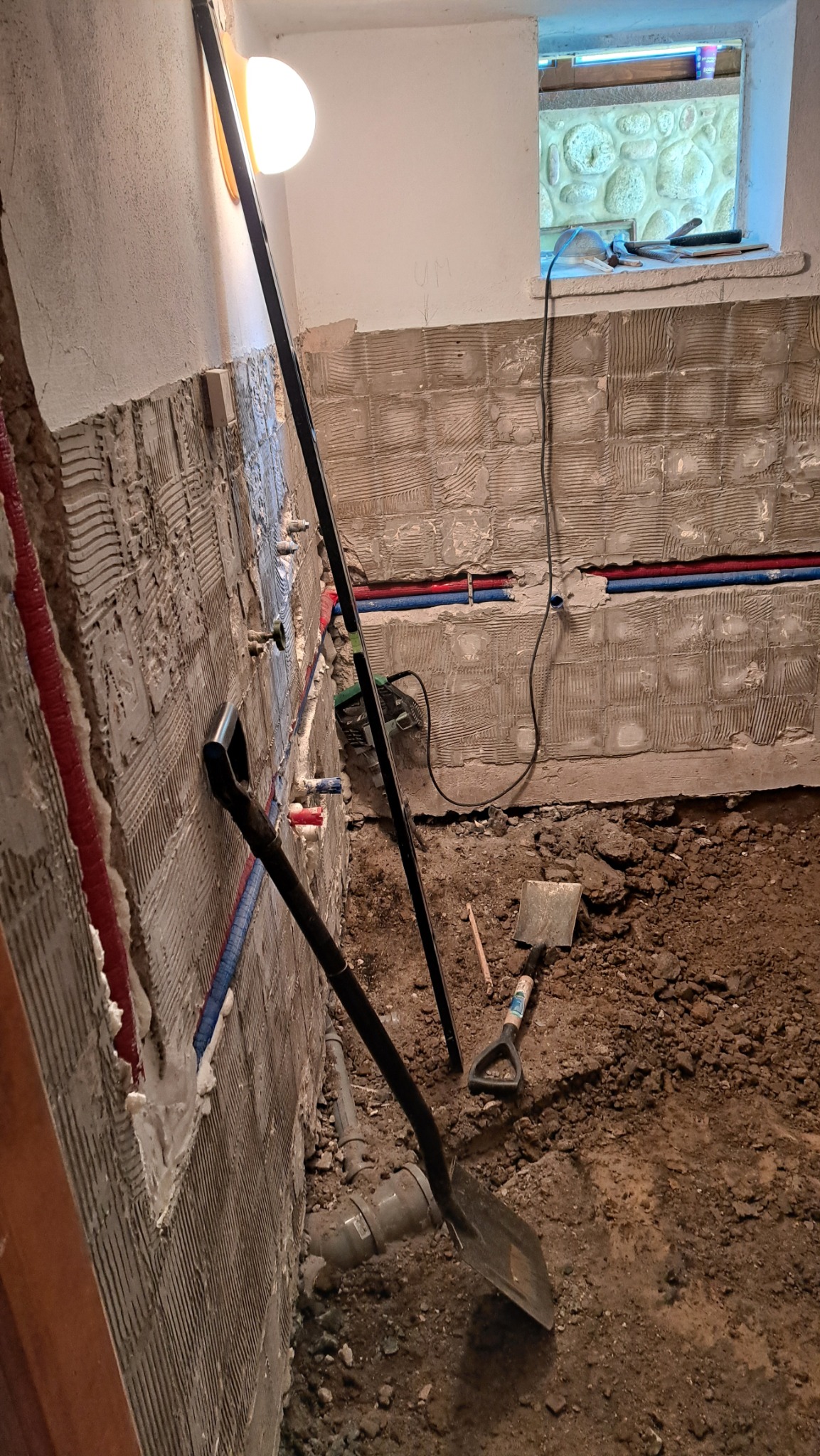 Przygotowanie do remontu: ściany z resztkami zaprawy, rury instalacyjne, łopata i ziemia na podłodze. Widoczne prace instalacyjne w pomieszczeniu.