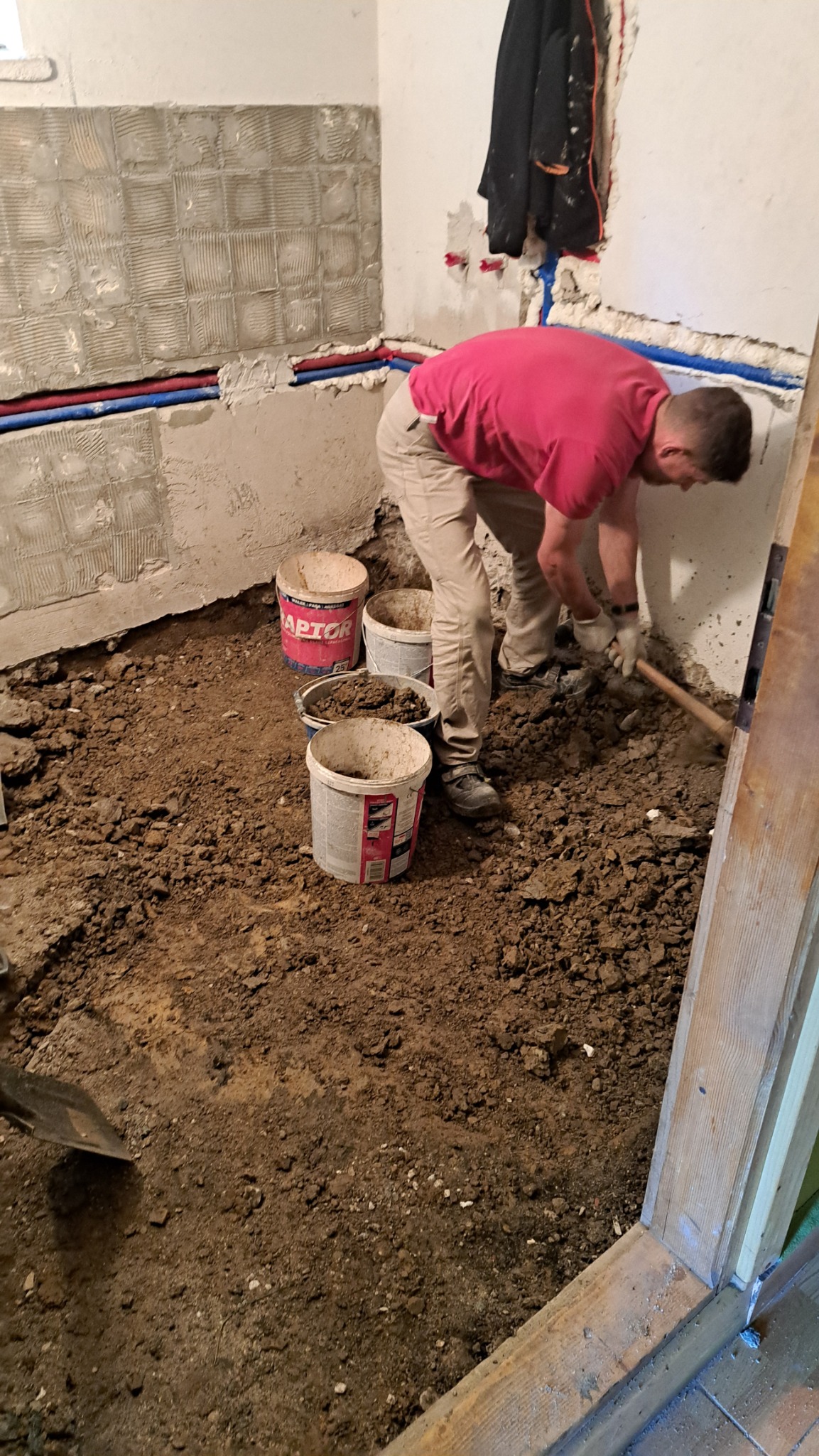 Demontaż posadzki w łazience: mężczyzna w czerwonej koszulce usuwa ziemię łopatą, widoczne ściany z pozostałościami płytek i rury instalacji.