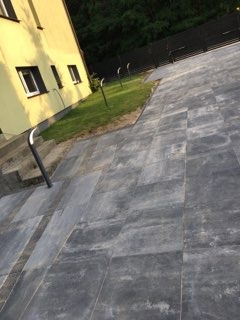 Ułożona, szara nawierzchnia z dużych płyt betonowych prowadząca od schodów domu do bramy wjazdowej, z widocznym fragmentem trawnika i lampami ogrodowymi.