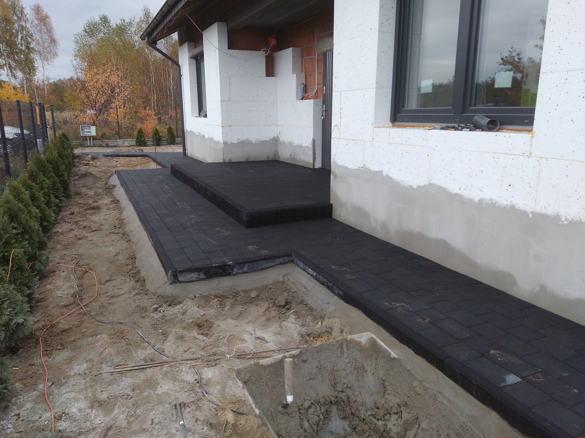 Ciemna kostka ułożona jako ścieżka i schody przed domem w trakcie prac wykończeniowych, widoczne przygotowane podłoże i wykop.