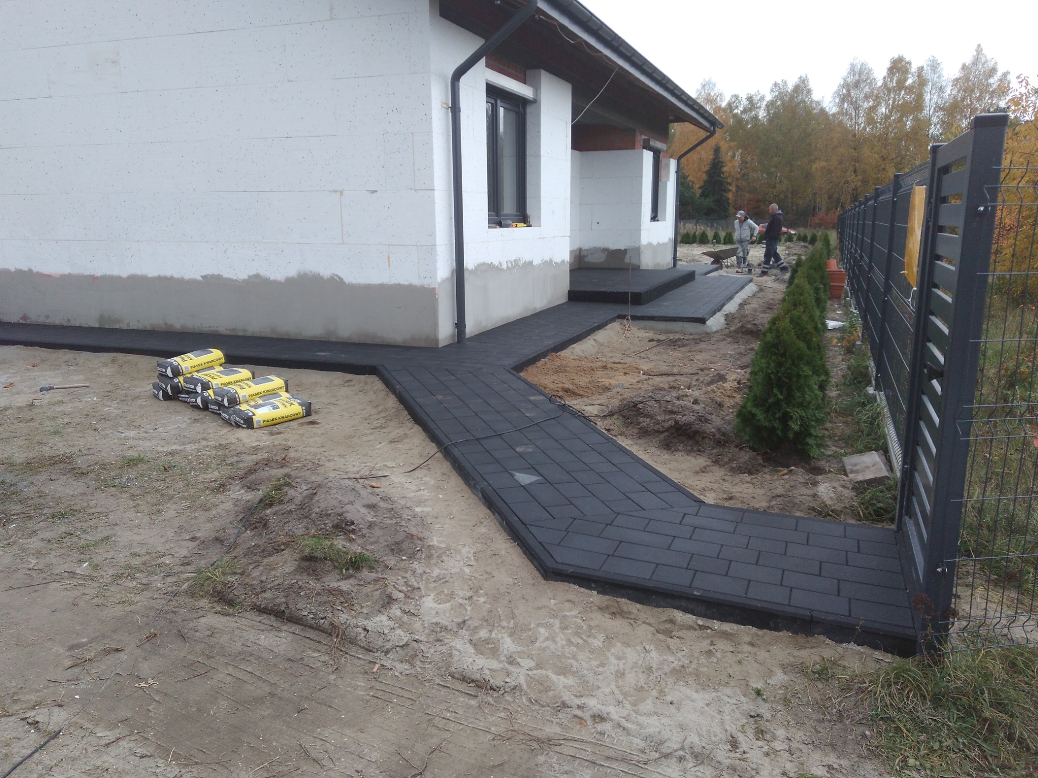 Ciemna kostka brukowa ułożona jako ścieżka wokół domu w trakcie budowy, widoczne worki z piaskiem i narzędzia, dwóch pracowników w tle.