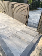 Nowoczesna brama wjazdowa z poziomymi panelami, otwierana automatycznie, prowadząca na podjazd wyłożony szarą kostką brukową z ciemnoszarym obramowaniem i wstawkami z jaśniejszej kostki. Widoczny...