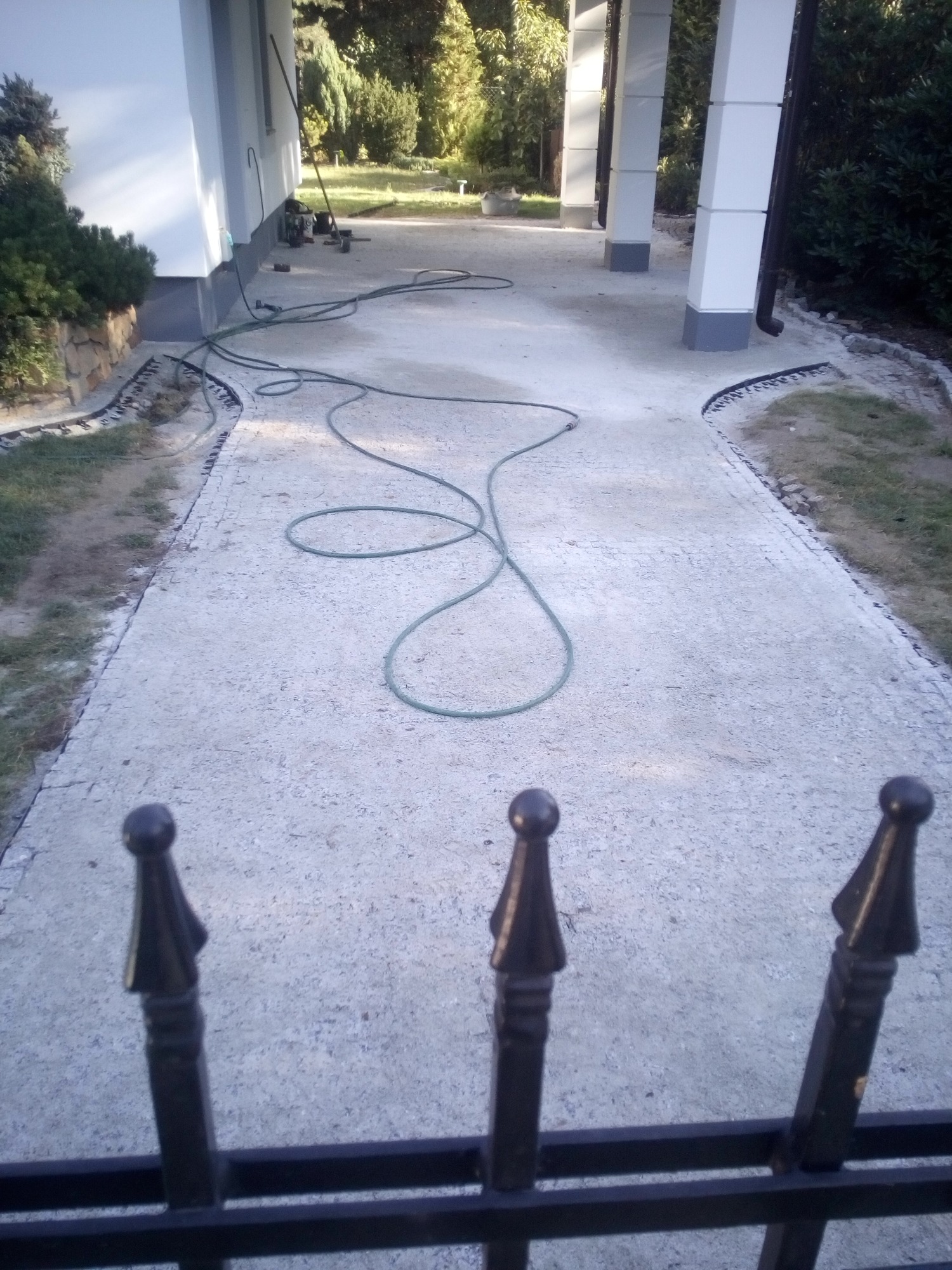 Przygotowanie podłoża pod ścieżkę z kostki, widoczny piasek, obrzeża i rozłożony wąż ogrodowy, ujęcie zza ozdobnego ogrodzenia.