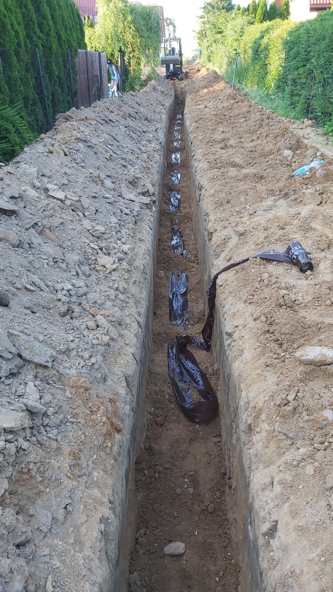 Długi, wąski wykop w ziemi z folią zabezpieczającą, widoczny z oddali mały traktor, prace ziemne w toku.