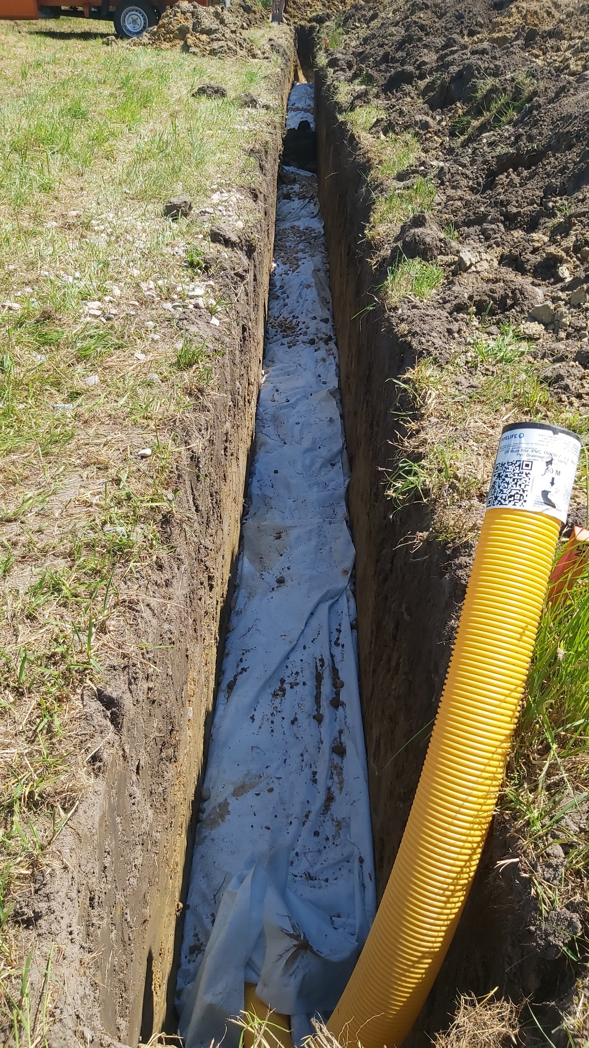 Wykop w ziemi wyłożony białą geowłókniną, obok leży żółta rura drenażowa karbowana z etykietą producenta.