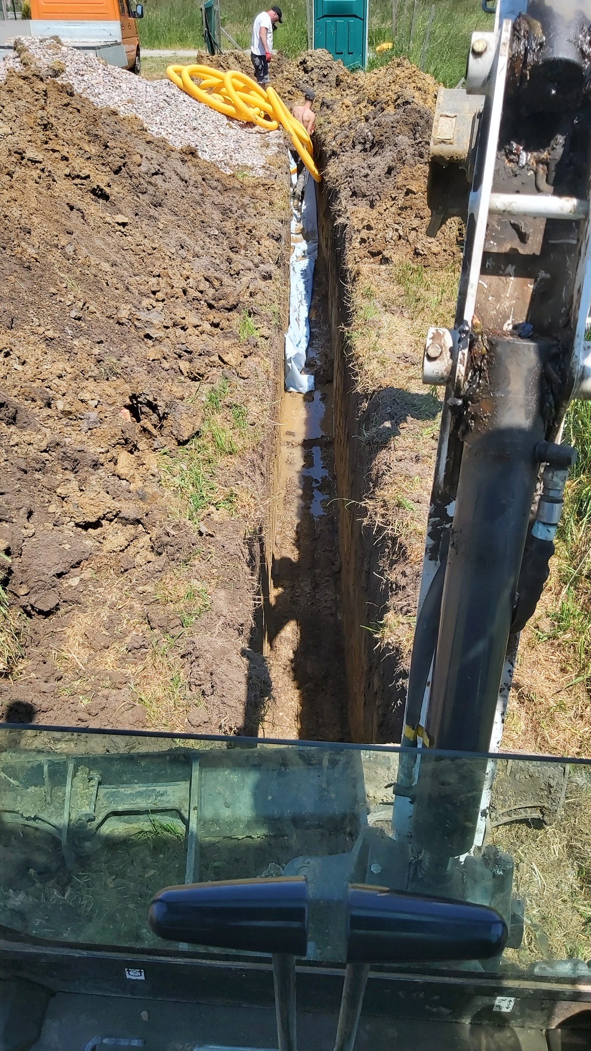 Wykopany wąski rów w ziemi, zabezpieczony białą geowłókniną, z żółtymi rurami na brzegu i widoczną częścią koparki z perspektywy operatora.