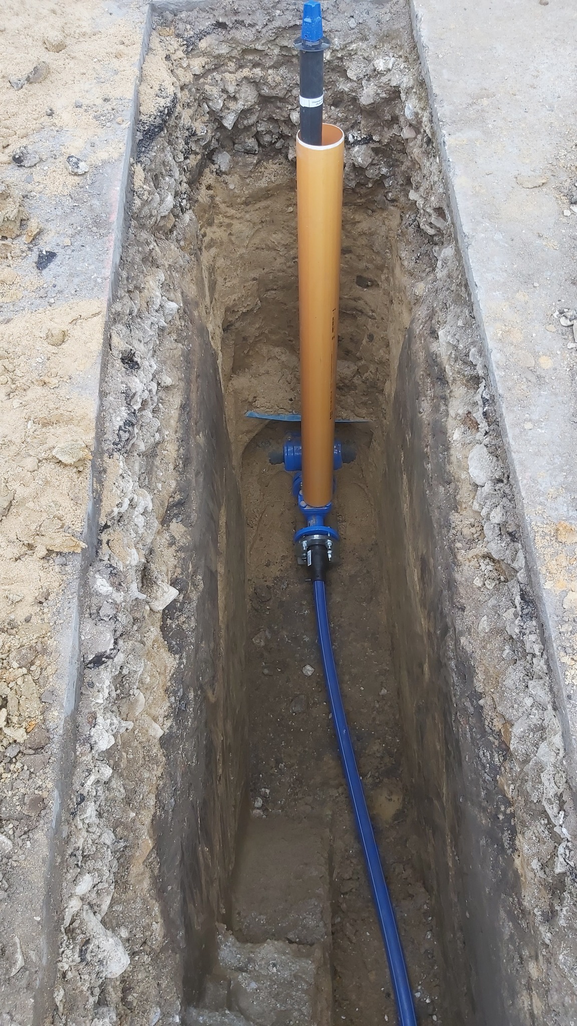 Wykop w ziemi z widoczną pionową rurą w kolorze piaskowym, połączoną niebieskimi elementami instalacji i niebieskim przewodem. Wokół wykopu widoczna ziemia z kamieniami.