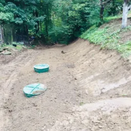 Dwa zielone włazy szamba na wykopanym terenie, widoczne ślady opon, w tle drzewa i zarośla.