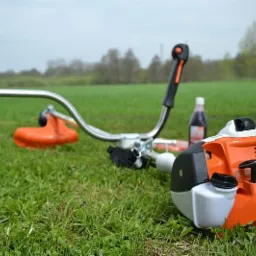Pomarańczowo-biała kosa spalinowa Stihl FS 56 leży na zielonej trawie, w tle butelka z płynem i drzewa.