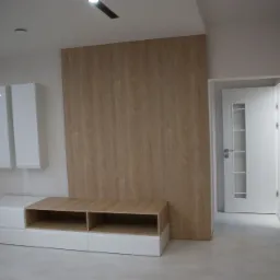 Nowoczesny salon z białymi szafkami wiszącymi i szafką RTV z drewnianym blatem, na tle ściany wyłożonej panelami imitującymi drewno, widok na białe drzwi.