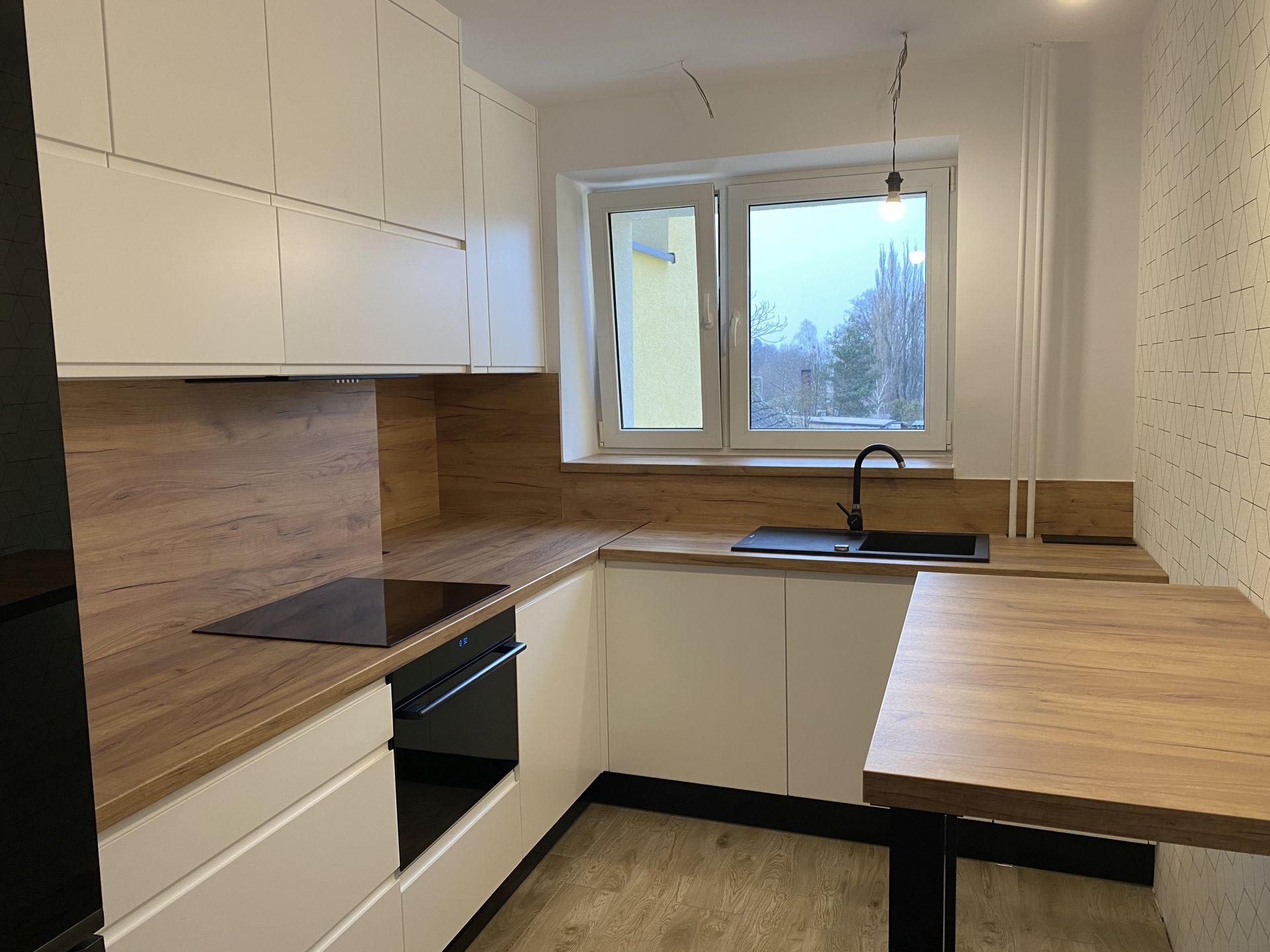 Nowoczesna kuchnia w odcieniach bieli i drewna z czarnymi akcentami, widok na okno z widokiem na drzewa, minimalistyczny styl.