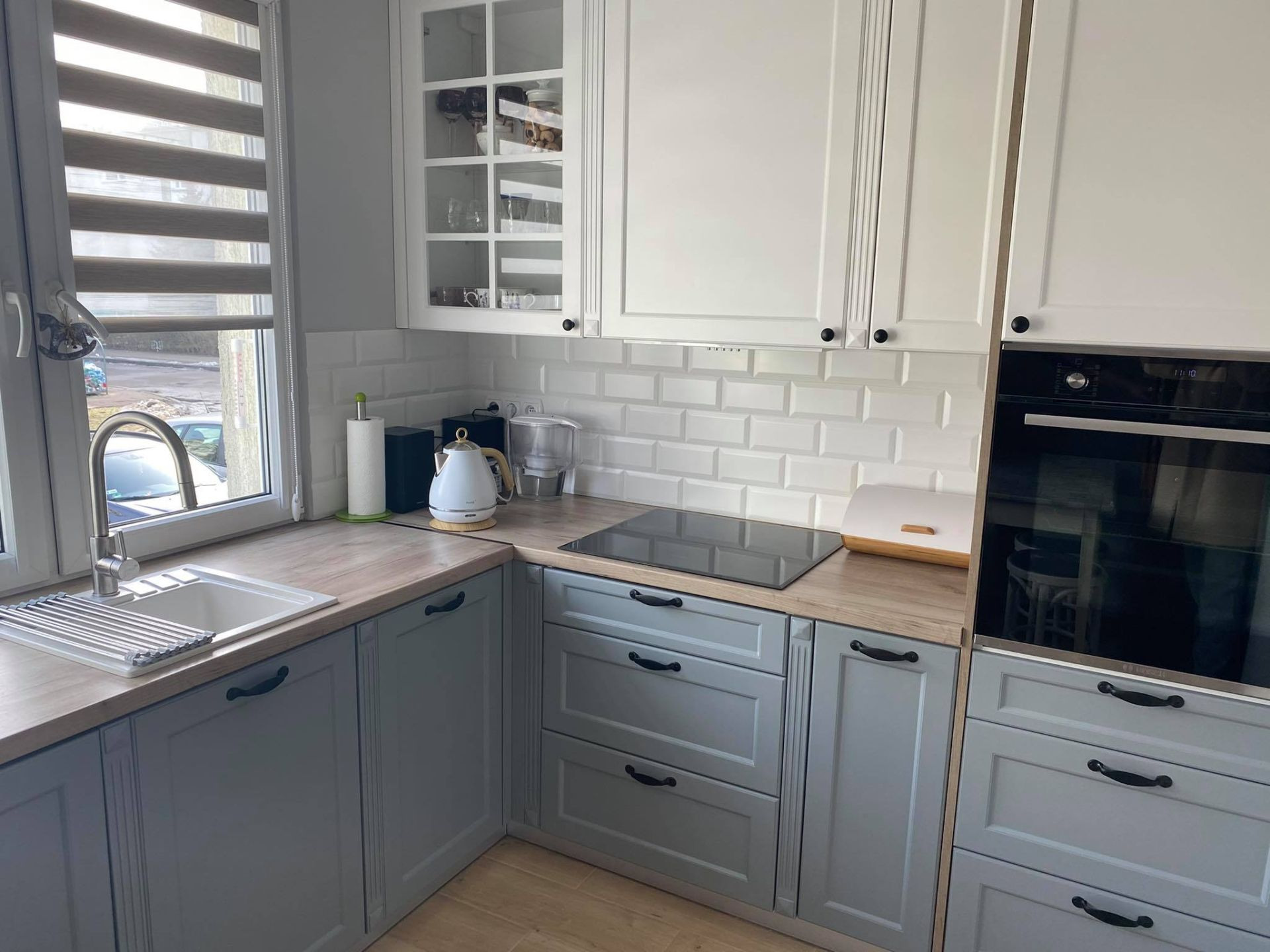 Jasna kuchnia z szarymi dolnymi szafkami i białymi górnymi, płytki typu metro na ścianie, drewniane blaty i widok na zewnątrz przez okno z roletami.