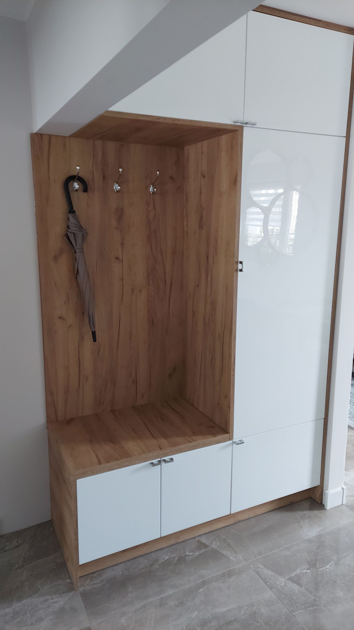 Wnęka garderoby z drewnianymi panelami i białymi szafkami, z szarym parasolem wiszącym na haczyku.