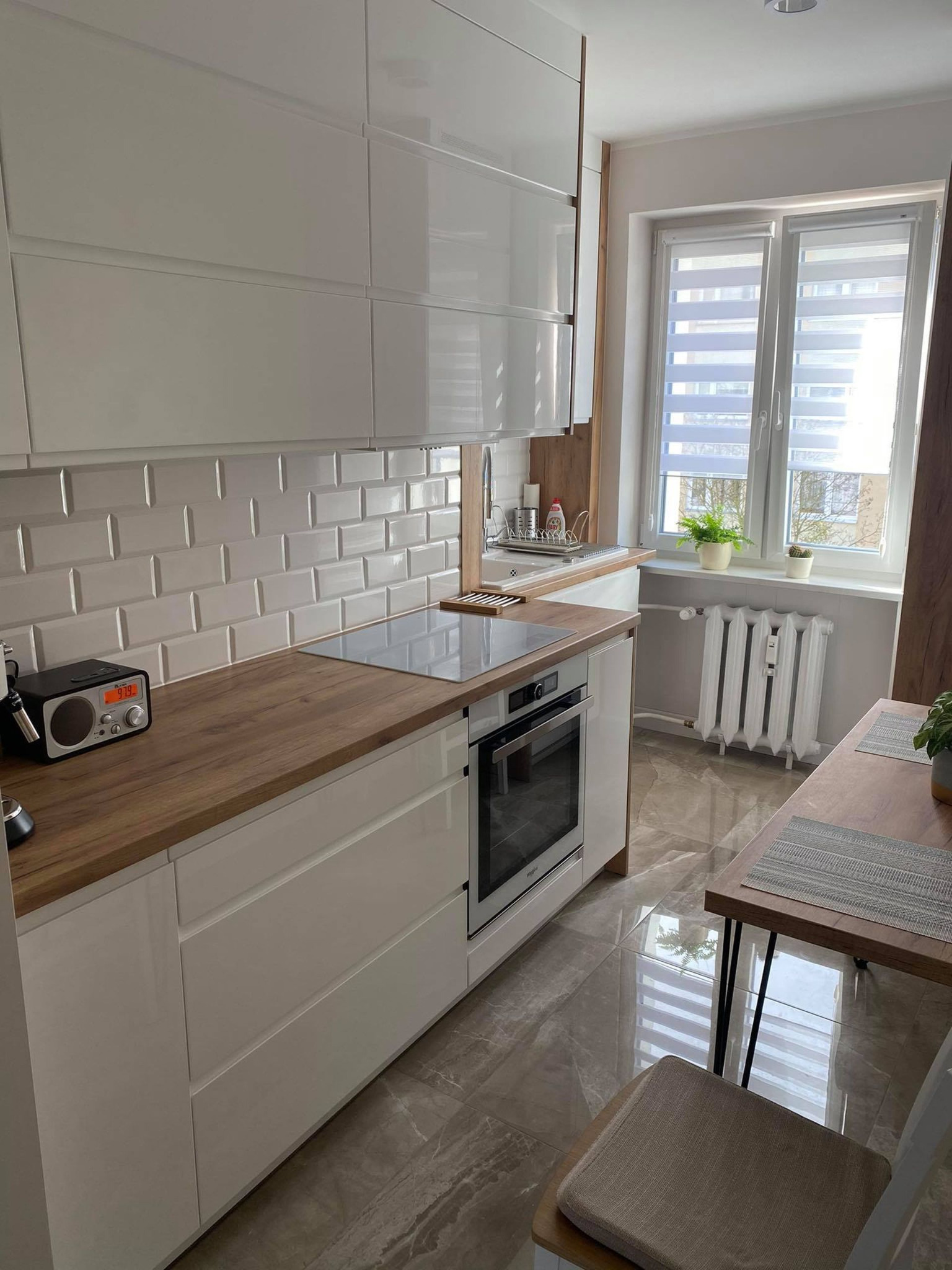 Nowoczesna kuchnia z białymi, lakierowanymi frontami, drewnianym blatem i płytkami typu metro, uzupełniona o radio retro na blacie i widok z okna na zieleń.